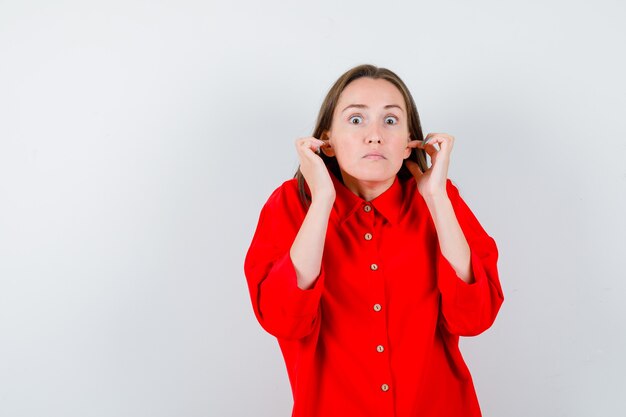 Portret młodej kobiety zatykającej uszy palcami w czerwonej bluzce i patrzącej na przestraszony widok z przodu