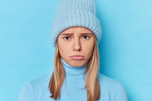 Portret młodej kobiety zdenerwowanej złymi wiadomościami, która żałuje, że zrobiła coś złego, ma nastrojowe wyrazy twarzy, nosi ciepły kapelusz i golf na białym tle na niebieskim tle. koncepcja negatywnych emocji