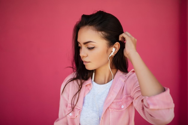 Bezpłatne zdjęcie portret młodej ładnej dziewczyny o ciemnych włosach, stojącej i słuchającej muzyki w słuchawkach, podczas gdy starannie odsunięte na bok na różowym tle