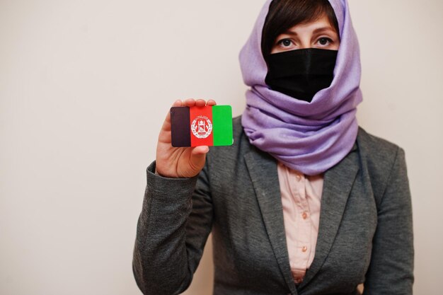 Portret młodej muzułmańskiej kobiety w formalnym stroju chroni maskę i hidżab na głowę trzymaj kartę flagi Afganistanu na na białym tle Koncepcja kraju koronawirusa