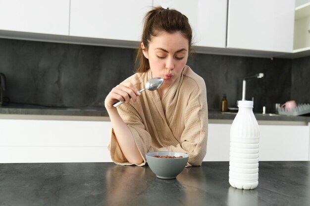 Portret młodej pięknej kobiety w szlafroku jedzącej płatki na śniadanie, opiera się na blacie kuchennym