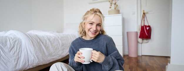 Portret młodej pięknej kobiety w zwykłych ubraniach siedzącej na podłodze sypialni z filiżanką herbaty