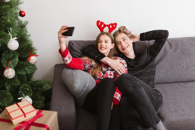 Portret młodej pięknej pary siedzącej na kanapie w domu i szczęśliwie biorącej selfie wraz z choinką w pobliżu