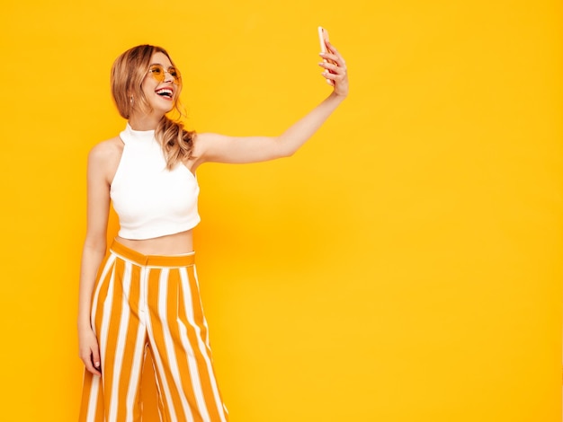 Portret młodej pięknej uśmiechniętej blond kobiety w modnych letnich ubraniach beztroska kobieta pozuje w pobliżu żółtej ściany w studio Pozytywny model zabawy w pomieszczeniu Wesoły i szczęśliwy Robienie zdjęć selfie