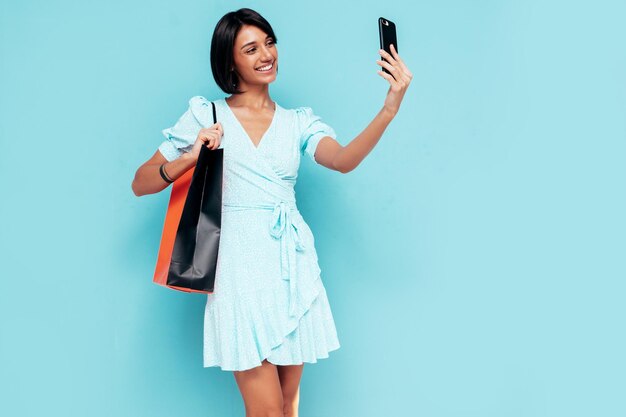 Portret młodej pięknej uśmiechniętej kobiety w modnej letniej sukience Beztroska kobieta pozuje w pobliżu niebieskiej ściany w studio Pozytywny model trzymający torbę na zakupy Wesoły i szczęśliwy Robienie selfie
