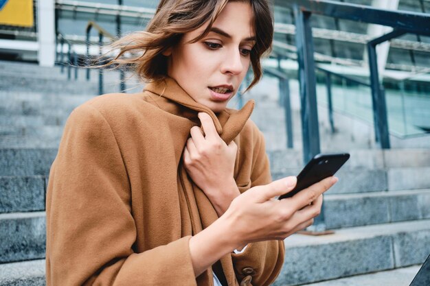 Portret młodej poważnej bizneswoman zapinającej płaszcz z zimna przy użyciu telefonu komórkowego na zewnątrz
