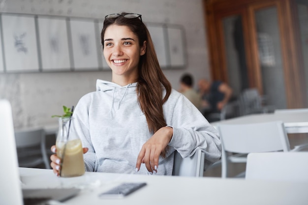Portret młodej studentki administracji biznesu śmiejącej się z problemów z trzecim światem podczas picia lemoniady za pomocą laptopa, aby nawiązać połączenie