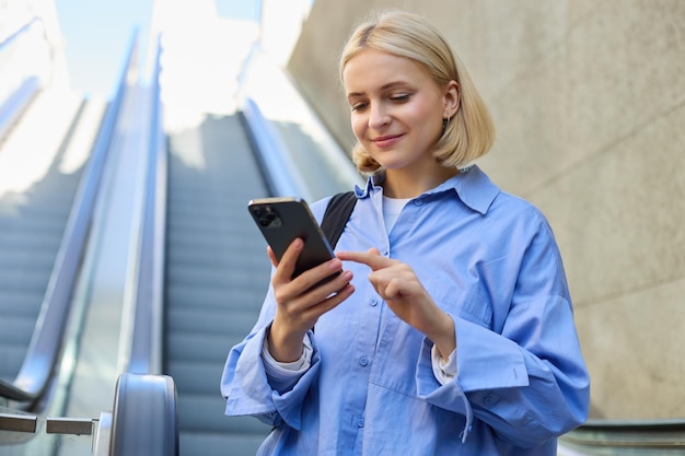 Bezpłatne zdjęcie portret młodej stylowej kobiety, pracownicy stojącej w pobliżu schodów ruchomych za pomocą telefonu komórkowego