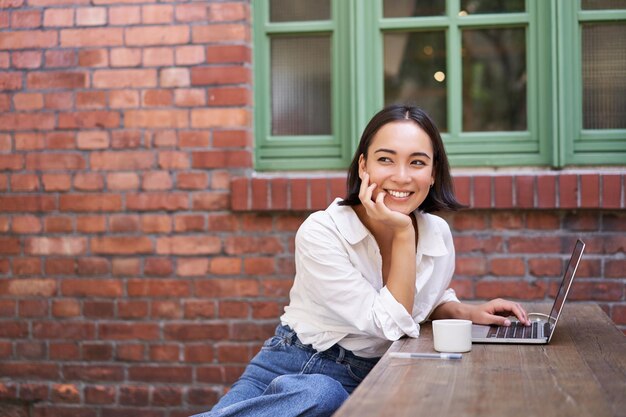 Portret młodej stylowej kobiety wpływowej siedzącej w kawiarni z filiżanką kawy i uśmiechającym się laptopem i