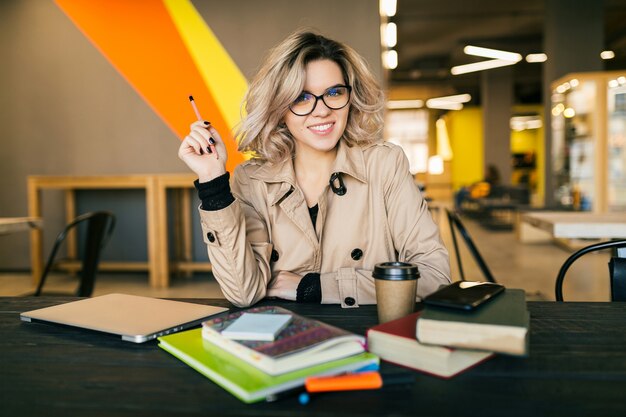 Portret młodej stylowej kobiety z pomysłem, siedząca przy stole w trenczu, pracująca na laptopie w biurze coworkingowym, w okularach, uśmiechnięta, szczęśliwa, pozytywna, zajęta