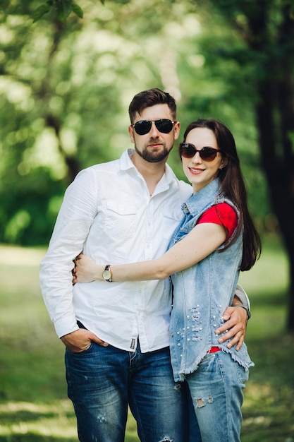 Portret młodej szczęśliwej pary zakochanej uśmiechniętej i obejmującej się w ogrodzie Słodcy kochankowie w stylowych ciemnych okularach przeciwsłonecznych i swobodnych ubraniach pozowanie i patrząc na kamerę w ciągu dnia w letnim parku