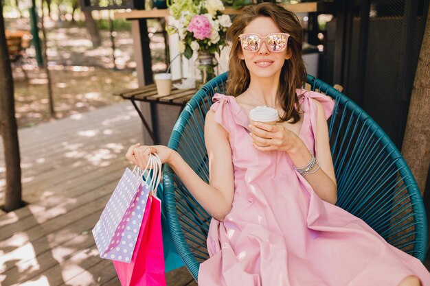 Portret młodej uśmiechniętej szczęśliwej atrakcyjnej kobiety siedzącej w kawiarni z torbami na zakupy pijącej kawę, letni strój modowy, różowa bawełniana sukienka, modna odzież