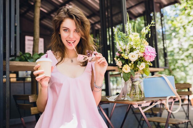 Portret młodej uśmiechniętej szczęśliwej ładnej kobiety siedzącej w kawiarni pijąc kawę, letni strój moda, różowa bawełniana sukienka, modne dodatki odzieżowe