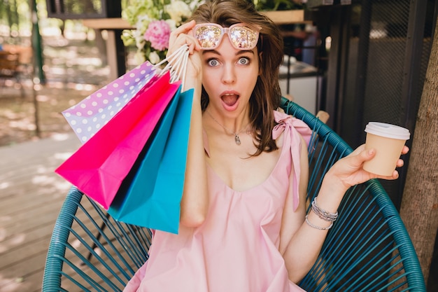 Portret młodej uśmiechniętej szczęśliwej ładnej kobiety z zaskoczonym wyrazem twarzy siedzi w kawiarni z torbami na zakupy pijąc kawę, letni strój mody, różowa bawełniana sukienka, modna odzież