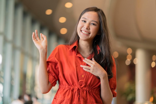 Portret młodej wesołej azjatyckiej uśmiechniętej kobiety gest ręki wskazuje copyspace stojącej w centrum handlowym z niebieskim światłem bokeh w tle uśmiechnięta azjatycka kobieta przypadkowa tkanina uśmiechnięta kamera spojrzenie