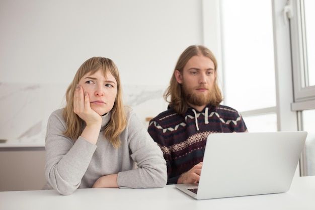 Bezpłatne zdjęcie portret młodej zdenerwowanej kobiety smutno patrzącej na bok, podczas gdy młody mężczyzna siedzi w pobliżu i pracuje na laptopie w domu
