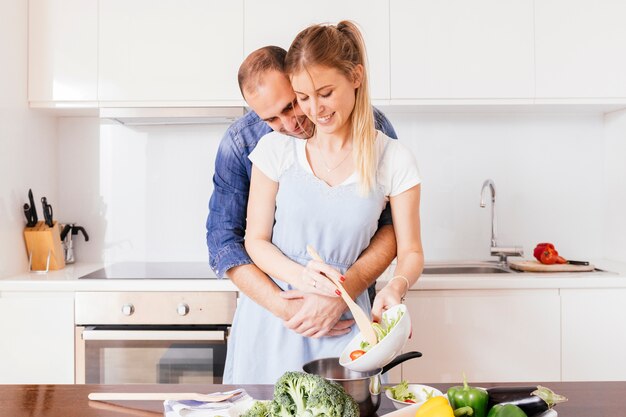 Portret młody człowiek obejmuje jego dziewczyny przygotowywa sałatki w kuchni