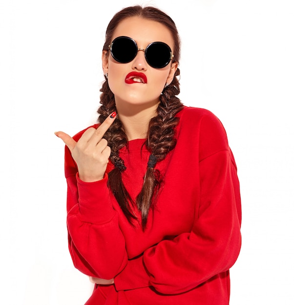 Portret młody szczęśliwy uśmiechnięty kobieta model z jaskrawym makeup i kolorowymi wargami z dwa warkoczami i okularami przeciwsłonecznymi w lato czerwieni ubraniach odizolowywających. pokazując kurwa znak