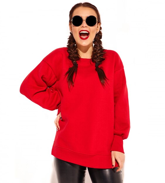 Portret młody szczęśliwy uśmiechnięty kobieta model z jaskrawym makeup i kolorowymi wargami z dwa warkoczami i okularami przeciwsłonecznymi w lato czerwieni ubraniach odizolowywających.