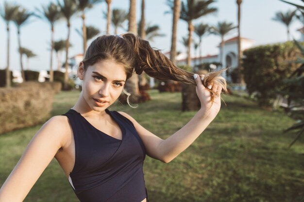 Portret niesamowita atrakcyjna młoda kobieta w odzieży sportowej bawi się długimi kręconymi włosami w parku tropikalnego miasta. Wyglądający, modny szczupły model, wakacyjny, figlarny, outfit