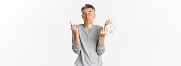 Portret nieświadomego mężczyzny w średnim wieku w okularach, wzruszającego ramionami, trzymając kartę kredytową z pieniędzmi stojącymi niezdecydowanie na białym tle