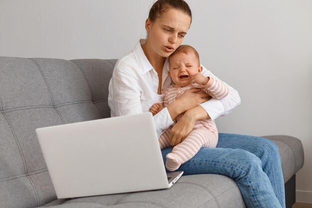 Portret nieszczęśliwej smutnej kobiety z kok włosy w białej koszuli i dżinsach, siedzącej na kaszel z płaczącą córeczką, próbującą uspokoić niemowlę, przytulając dziecko i uciszając.