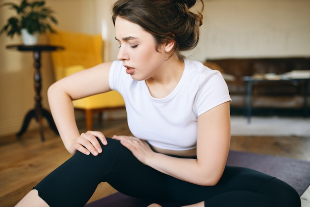 Portret nieszczęśliwej wysportowanej dziewczyny chwytającej kolano, próbującej masować obolałe okolice, niezdolnej do uprawiania jogi z powodu kontuzji sportowej, odczuwającej ból.