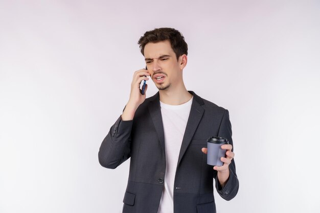 Portret niezadowolony przystojny biznesmen rozmawia przez telefon komórkowy i trzyma gorącą kawę na białym tle nad białym tłem