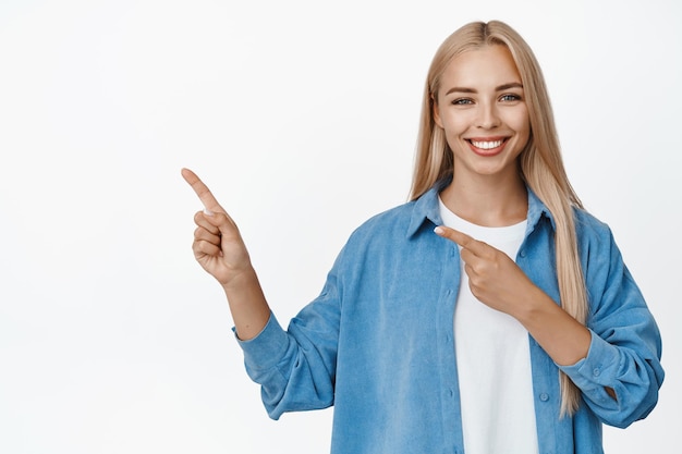 Portret nowoczesnej blond kobiety wskazując palcami w lewym górnym rogu, uśmiechając się do kamery pokazującej sprzedaż baner reklamowy stojący na białym tle