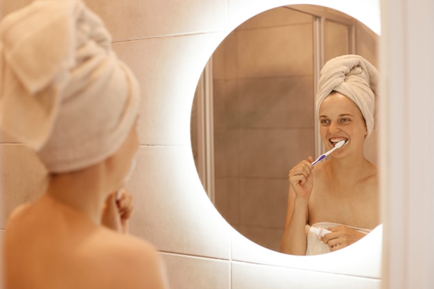 Bezpłatne zdjęcie portret optymistycznej ujmującej kobiety, która myje zęby, ma procedury higieniczne po wzięciu prysznica w łazience, stojąc z białym ręcznikiem na włosach.
