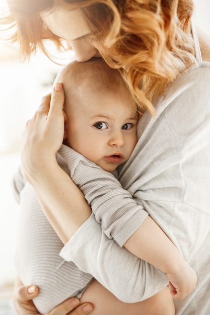 Portret patrzeje kamerę z zainteresowanym wyrażeniem słodki małe dziecko podczas gdy matka czule tuli palming i całuje jej nowonarodzonego dziecka.