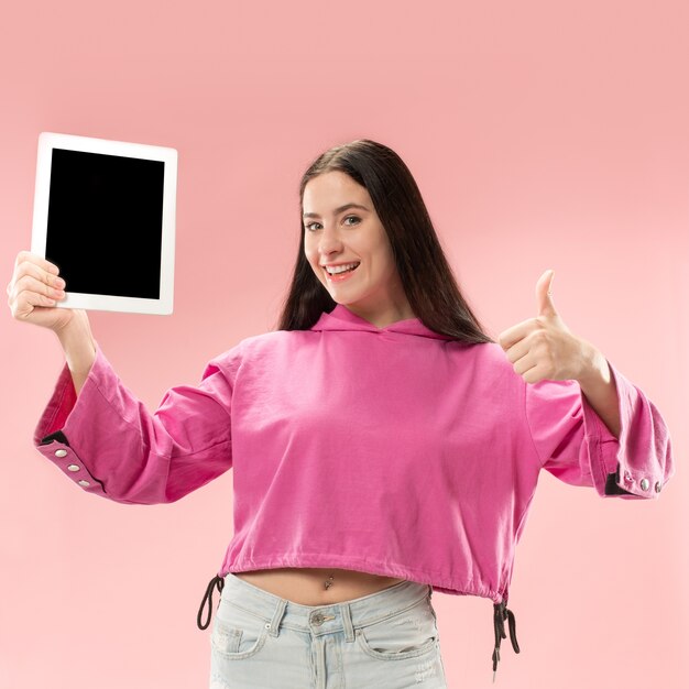 Portret pewnej dziewczyny dorywczo pokazując pusty ekran laptopa na białym tle nad różowym tłem.
