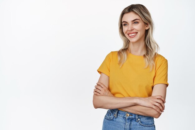 Portret pewnej siebie blond dziewczyna uśmiecha się skrzyżowanymi ramionami na klatce piersiowej i patrzy na bok stojąc w żółtej koszulce i dżinsach na białym tle