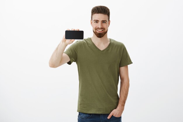 Portret pewnie przystojny brodaty mężczyzna współpracownik trzymający poziomo smartfon i uśmiechnięty zachwycony
