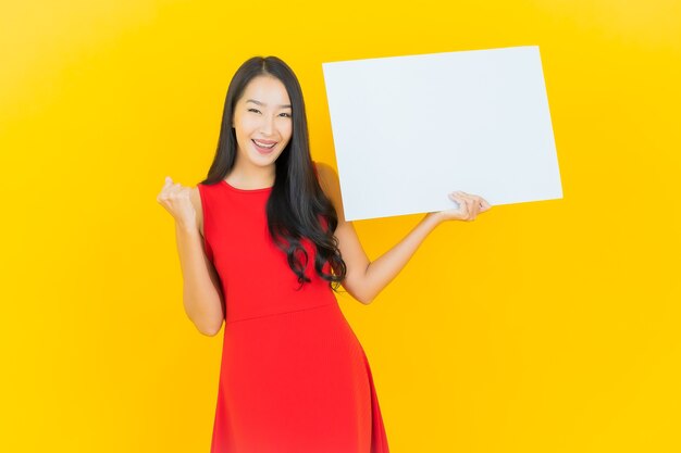 Portret piękna młoda azjatycka kobieta uśmiech z pustą białą tablicą na żółtej ścianie
