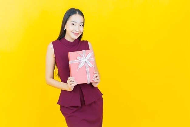 Portret piękna młoda azjatycka kobieta uśmiecha się z czerwonym pudełkiem na żółtej ścianie