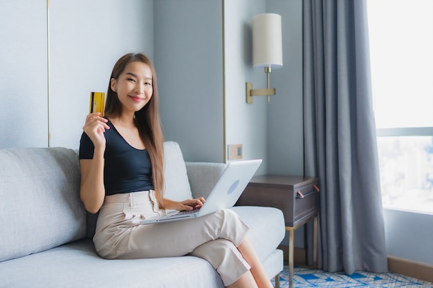 Portret piękna młoda azjatycka kobieta używa inteligentnego telefonu komórkowego lub laptopa z kartą kredytową na kanapie w salonie