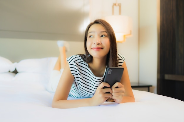 Portret piękna młoda azjatycka kobieta używa inteligentnego telefonu komórkowego na łóżku we wnętrzu sypialni