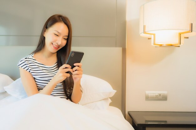Portret piękna młoda azjatycka kobieta używa inteligentnego telefonu komórkowego na łóżku we wnętrzu sypialni