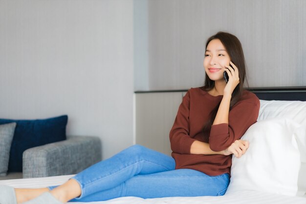 Portret piękna młoda azjatycka kobieta używa inteligentnego telefonu komórkowego na łóżku we wnętrzu sypialni