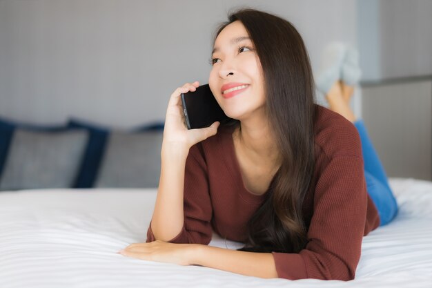 Portret piękna młoda azjatycka kobieta używa inteligentnego telefonu komórkowego na łóżku we wnętrzu sypialni