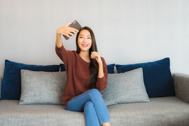 Portret piękna młoda azjatycka kobieta używa inteligentnego telefonu na kanapie we wnętrzu salonu