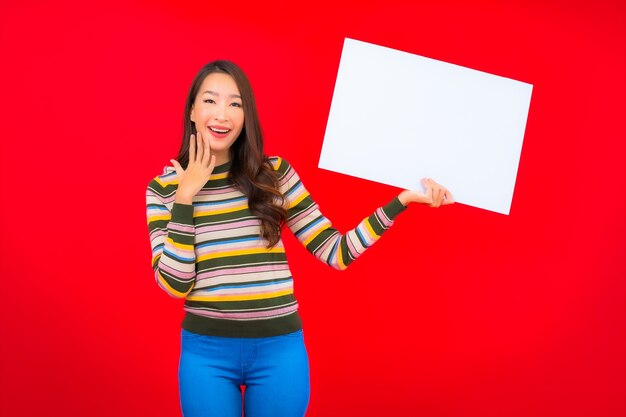 Portret piękna młoda azjatycka kobieta z białym pustym billboardem na czerwonej ścianie