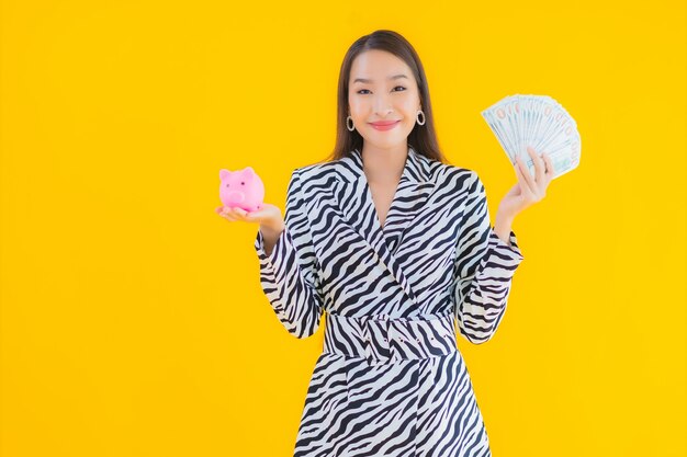 Portret piękna młoda azjatycka kobieta z piggy bank i gotówką lub pieniądze na żółto