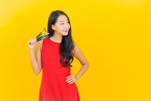 Portret piękna młoda azjatykcia kobieta z kosmetycznym pędzelkiem na żółtej ścianie