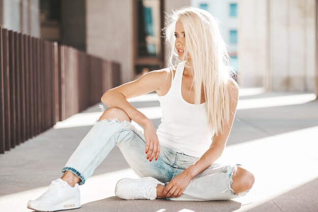 Portret piękna śliczna blond dziewczyna w białej koszulce i cajgach pozuje outdoors. Śliczny dziewczyny obsiadanie na asfalcie na ulicie