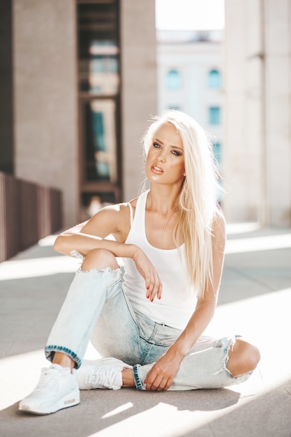 Portret piękna śliczna blond dziewczyna w białej koszulce i cajgach pozuje outdoors. Śliczny dziewczyny obsiadanie na asfalcie na ulicie