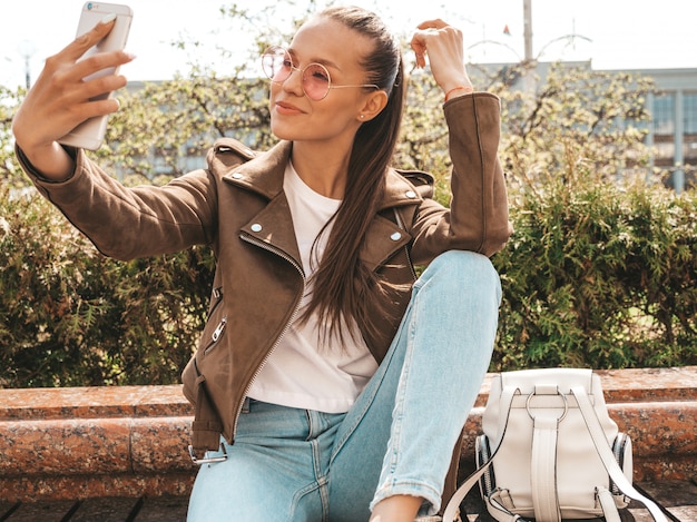 Portret piękna uśmiechnięta brunetki dziewczyna w lato modnisia kurtce i cajgach Wzorcowy bierze selfie na smartphone