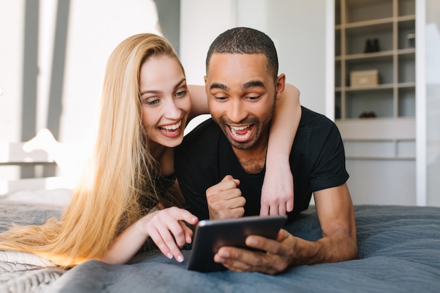 Portret Piękne Chwile Szczęśliwej Pary ślicznej Młodej Kobiety Z Długimi Blond Włosami I Przystojnego Faceta, Surfując W Internecie Na łóżku W Nowoczesnym Mieszkaniu. Dobra Zabawa, Młoda Rodzina, Miłość