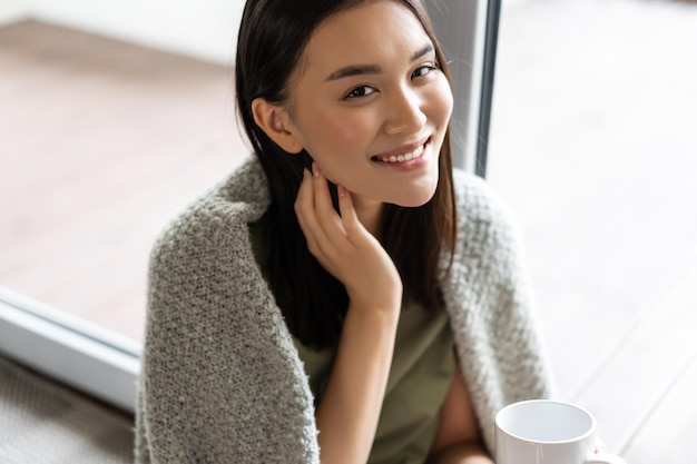 Portret pięknej azjatyckiej kobiety owiniętej w koc, pijącej kawę i uśmiechającej się, relaksującej się w domu na...